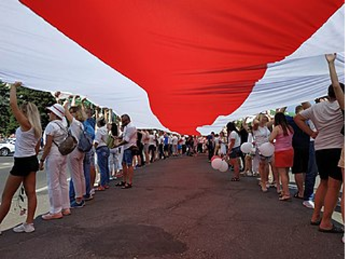 People lining both sides of a road, collectively holding a red and white striped flag above their heads.