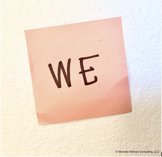 A coral colored sticky note on a white wall that has the word ‘WE’ written on it with black ink.