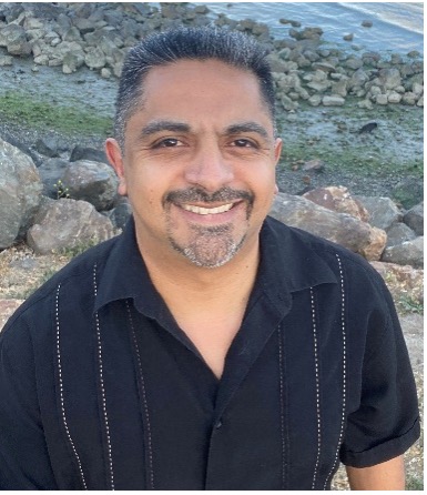 A person with short hair, a mustache, and a goatee, smiling and wearing a black button-up, short-sleeved shirt, standing in front of a rocky coast.