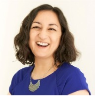 Image of a person, who has shoulder-length curly hair and is wearing a blue top and dissent-collar-style necklace, laughing.