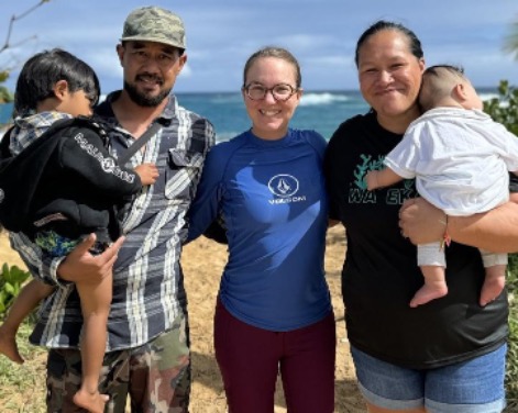 Three people with arms around each other's waists stand on a sandy beach with an ocean in the background. On the left is a man carrying a toddler, in the middle is a woman wearing glasses, and on the right is a woman carrying a baby.