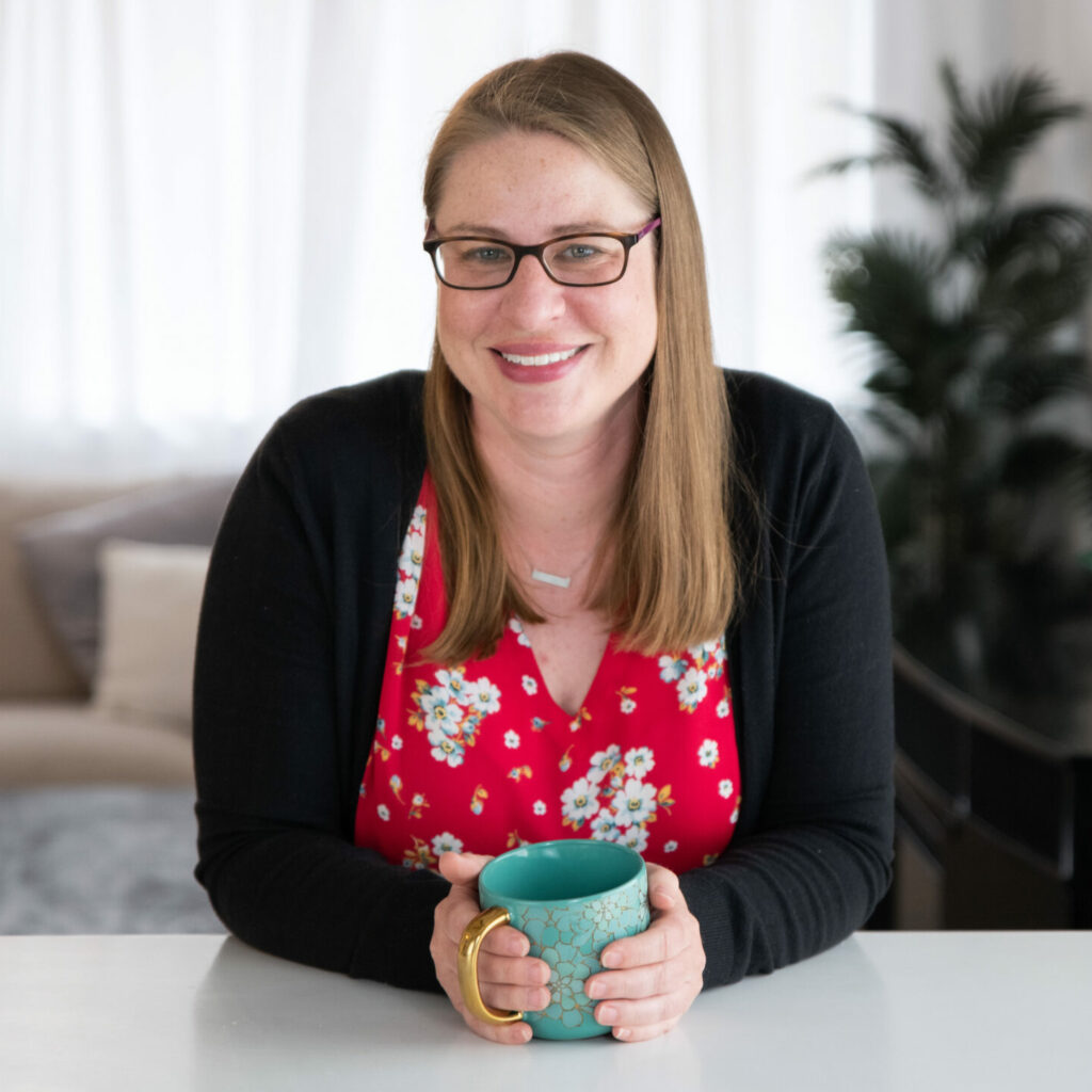 Elizabeth Grim, a white woman with long blond hair and glasses, sits at a table holding a mug.